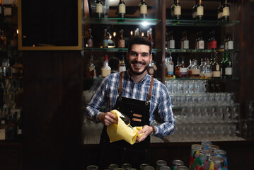 Young happy bartender smiling and polishing drinking glass in the cafeteria or bar with retro wooden look. Man cleaning glass with rug behind the counter.