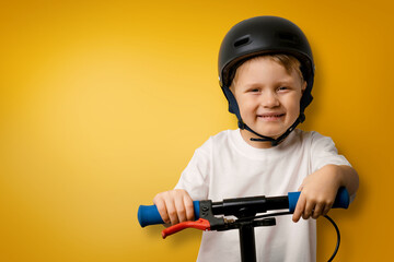 young smiling boy with helmet standing on kick scooter on yellow background with copy space