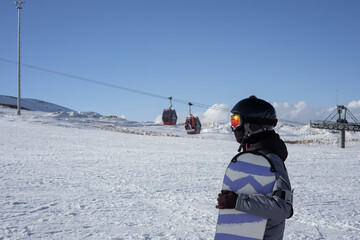 Girl or boy in ski helmet, sunscreen mask and balaclava with snowboard stands against the backdrop of snow-covered mountain ski slope and a cloudy sky. Winter. Sport and travel content  