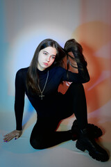 A young female fashion model with dark, long hair. A girl in dark clothes, with thick-soled shoes sits on a light background.