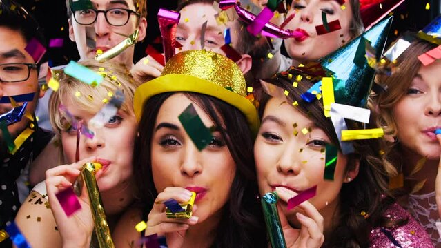 Party blower, confetti and group of people enjoying time together. Holiday event, celebration and happy friends blowing birthday whistle, having fun with hats and partying on dark studio background.