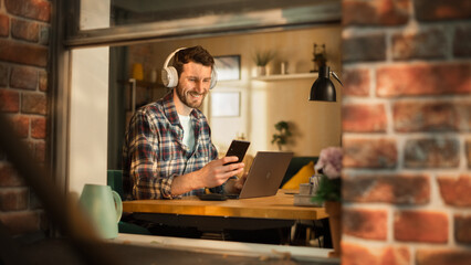 Handsome Caucasian Man Working on Laptop Computer while Sitting Behind His Desk at Home Office. Freelance Entrepreneur Listening to Music, Checking Smartphone. View From Outdoors into Window.