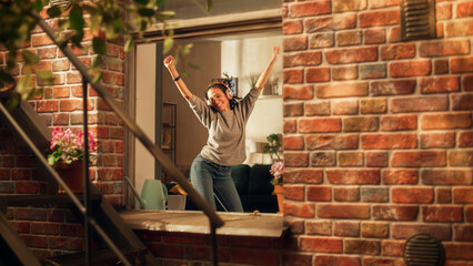 Latin Woman wearing Headphones and Dancing in her Apartment and Having Fun. Joyful Girl Feeling Happy and Carefree. Cheerful Female Is So Happy That you Want Join Her. Shot Throught Apartment Window. - obrazy, fototapety, plakaty