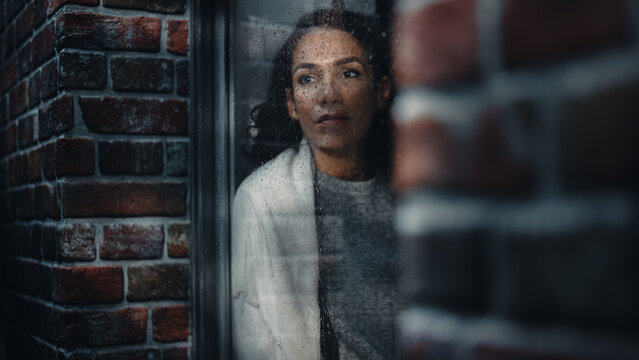 Melancholic Latin Woman Holding Cup of Coffee and Looking out of Window During Rainy Day. Female Thinking About Difficulties in Relationships with Boyfriend. Depression and Human Emotions.