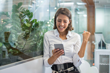 Excited cheerful young business woman with fist up receiving good news on smart phone and screaming...