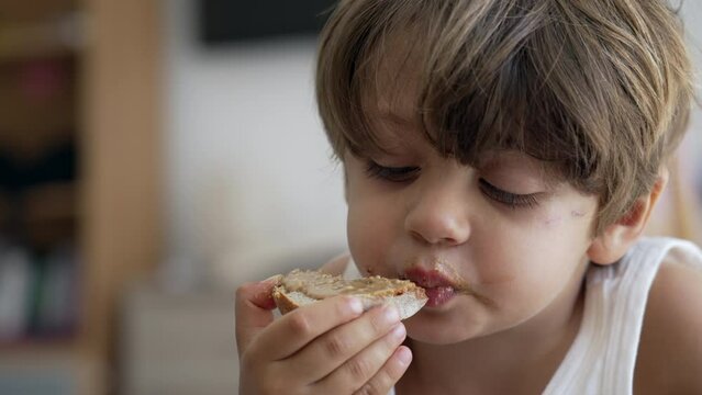 One Small Boy Eating Bread With Peanut Butter. Portrait Of A Child Taking A Bite Of Toast Carb Food In Morning Breakfast Or Snack