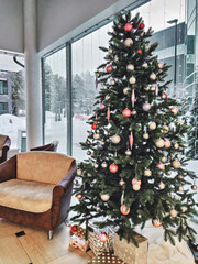 Christmas tree decorated with Christmas balls and garland, with gift boxes in the airy lobby beside the windows