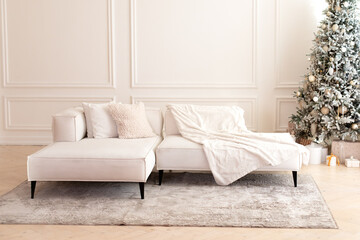 Comfortable couch with three cushions and a blanket standing in bright room interior with grey carpet in a modern interior. Modern interior living room with large light  corner sofa and Xmas tree.