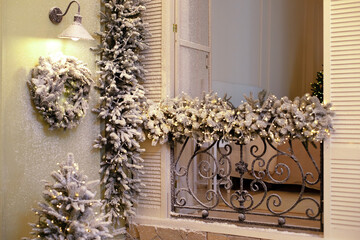 Snow covered balcony with wrought french iron railings with open shutters and doors. Old window with wrought-iron balcony decorated branches fir and glowing garland on Christmas
