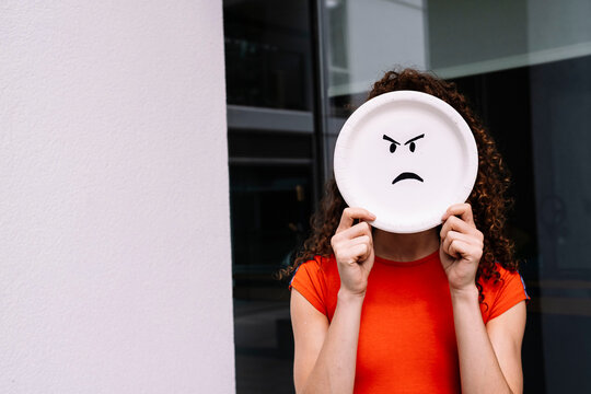 Young woman holding angry emoticon plate over face