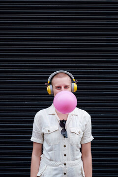 Non binary person blowing bubble gum wearing headphones listening to music in front of shutter