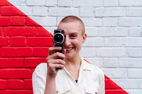 Non-binary person covering eye with video camera in front of two toned wall
