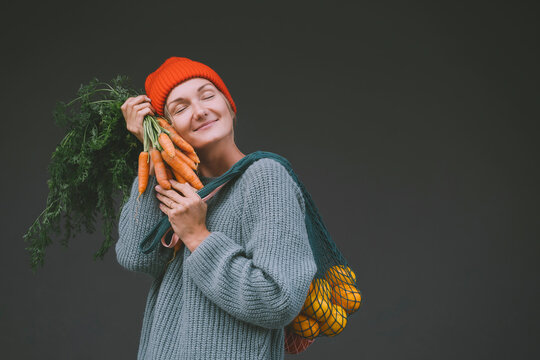 Smiling woman with eyes closed holding carrots and oranges in front of gray wall