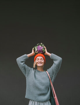 Smiling woman holding purple cauliflower on head in front of gray wall