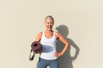 Happy woman with hand on hip in front of cream colored wall on sunny day