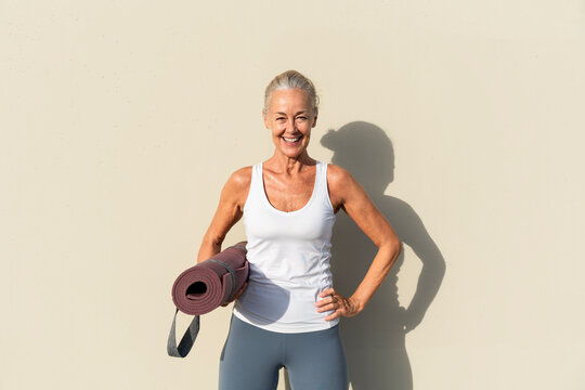 Smiling woman with yoga mat in front of cream colored wall