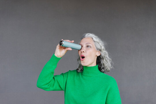 Surprised woman with mouth open looking through monocular in front of wall