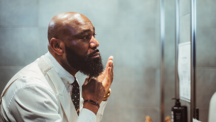 A profile view of a handsome bald black man grooming, brushing, and moisturizing the beard hair in...