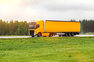 A yellow modern truck with a semi-trailer transports cargo in rainy weather against the backdrop of a forest. Delivery of goods by road transport, loistics. industry