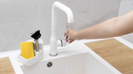 The girl opens a modern white water faucet in the kitchen. Detergent, sponge and dishwashing brush. Beautiful interior. Wall with white marble tiles.