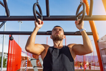 Man in a black t-shirt pulls up on gymnastic rings outdoors, exercising on the sports ground.