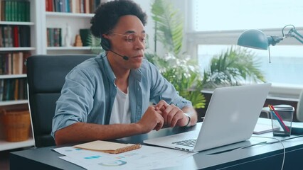 Young professional African American man consulting company specialist talking with client via video link in laptop using headphones with microphone and making notes in notebook sits at office desk