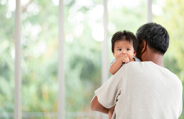Handsome father holding his little baby with black hairstyle on shoulder and looking through big window.