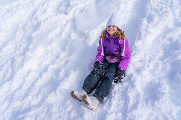 Fototapeta na wymiar A pretty girl in a purple ski suit slides down from the snow hill. Winter time children's games concept.