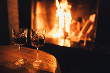 Two glasses of red wine on wooden table with cozy fireplace flame on the background.