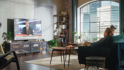Young Handsome Man Sitting in Relaxed Pose on a Sofa and Watching TV with Live News During the Day...