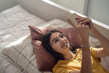 Top view of caucasian teenage girl browsing phone while lying on bed on her back