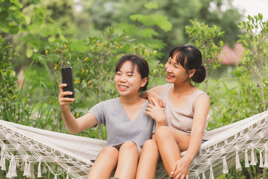 Young Asian teenager women sitting on hammock taking selfie outdoor rounded by green trees. Happy student friend enjoy vacation and taking picture together.