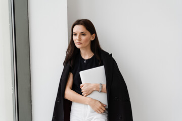 Confident young woman is standing with laptop on white background in the office. Attractive girl programmer is posing with laptop after remote work.