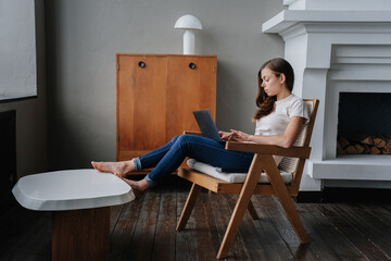 Disappointed Hispanic young woman in casual clothes sitting on chair with laptop at home against fireplace and cabinet. Caucasian attractive female entrepreneur remote working, using computer.