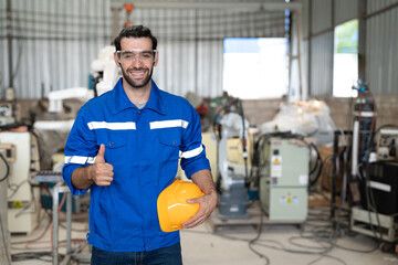 Portrait of male engineer in uniform smiling and showing thumbs up at industrial factory. Technician man standing holding helmet safety in manufacturing workshop.