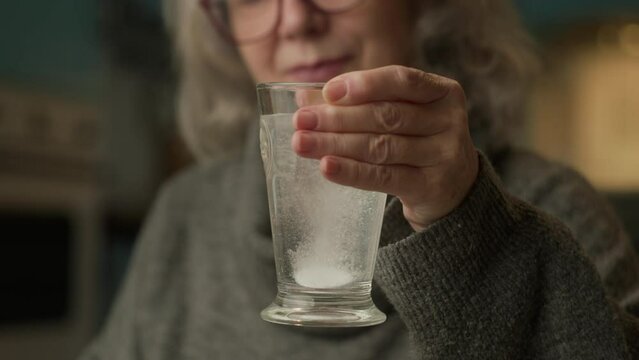 Elderly Hands Holding Glass Of Water With Effervescent Pill. Senior female taking medicine sitting at home.
