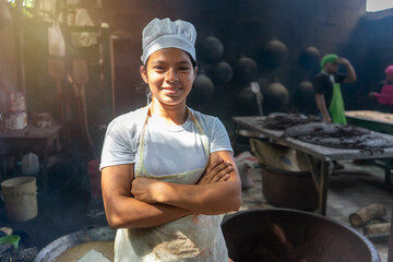 Young Latina woman from Nicaragua dressed in her cook uniform in a kitchen with traditional Latin American stoves and comales - obrazy, fototapety, plakaty
