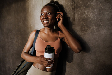 Young sporty smiling african woman in headphones holding bottle