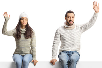 Young man and woman with winter jumpers sitting on a blank panel and waving