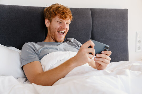 Young handsome enthusiastic redhead man in gray t-shirt playing mobile game