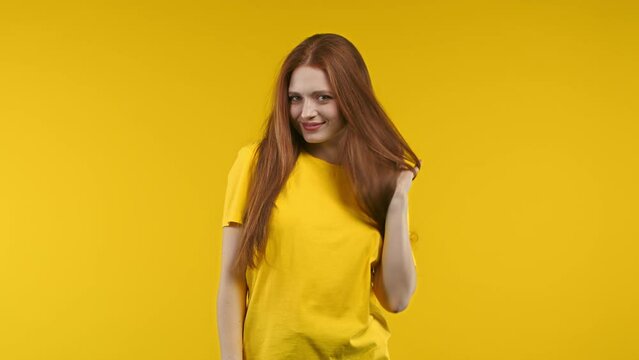 Playful ginger woman smiling, flirting to camera on yellow studio background. Cute trendy girl corrects her long awesome hair.