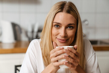 Blonde white woman smiling while drinking coffee in kitchen