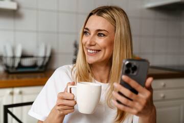 Blonde white woman drinking coffee and using cellphone in kitchen