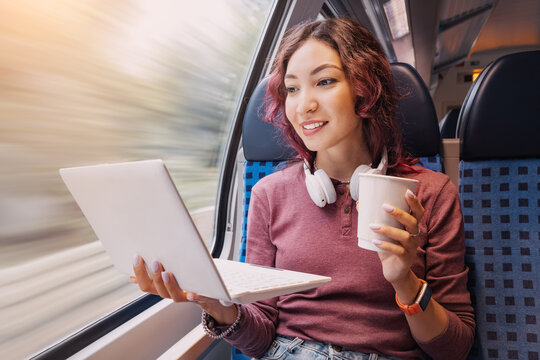 Happy young girl rides in a modern intercity train and remotely via Wi Fi Internet works or studies or watches a movie and drinking coffee
