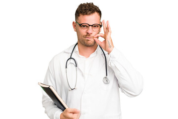 Young doctor caucasian man holding a book isolated with fingers on lips keeping a secret.