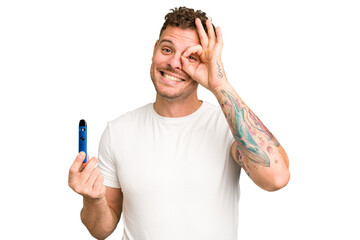 Young caucasian man holding a electronic cigarette isolated excited keeping ok gesture on eye.