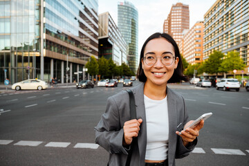 Young woman holding cellphone and smiling while standing at the city street