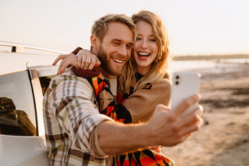Happy white couple taking selfie photo by car while walking at seashore