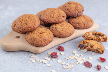 Oatmeal cookies on a wooden board, oatmeal and dried cranberries on a gray table. Healthy baking concept.