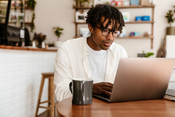 African man in glasses working on laptop and drinking coffee while sitting in cafe